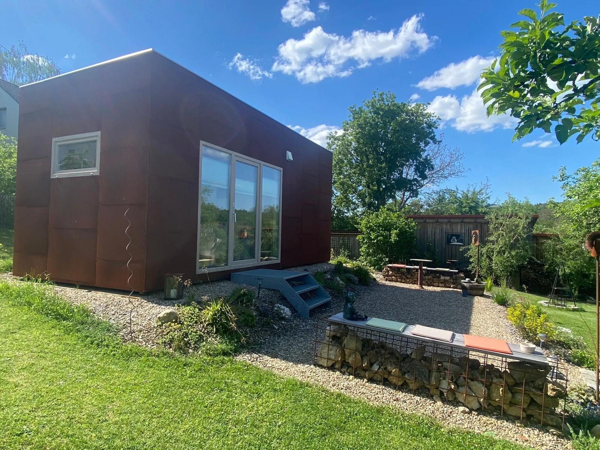 Das Tiny House von der Seite vor blauem Himmel, davor Kiesterrasse und Trockenmauer