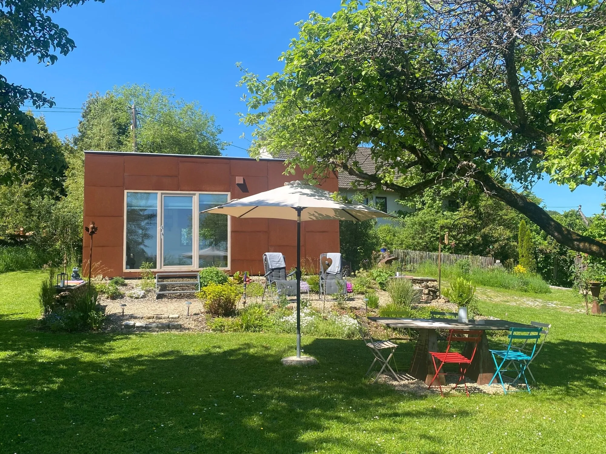 Das Tiny House im Obstgarten, davor Liegestühle und ein Tisch mit bunten Stühlen unter dem Sonnenschirm